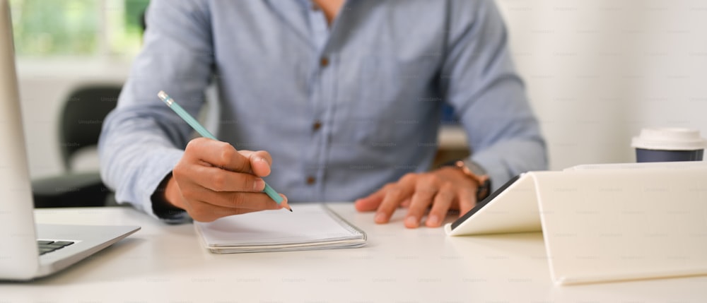 Abgeschnittene Aufnahme eines Geschäftsmannes, der einen Stift auf einem leeren Notizbuch am Büroschreibtisch hält.