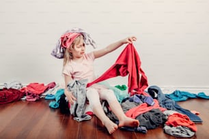 Kid playing with clothes on head. Cute Caucasian girl sorting clothes. Adorable funny child arranging organazing clothing. Messy stack of clothes things on floor. Home chores housework.