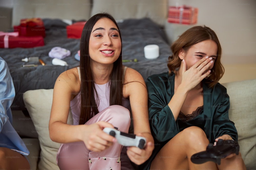Beautiful merry dark-haired gamer and her friend with joypads in their hands sitting on the floor
