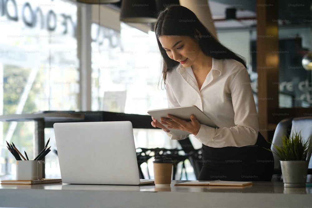 Jeune femme d’affaires confiante debout sur le lieu de travail et travaillant sur une tablette numérique.