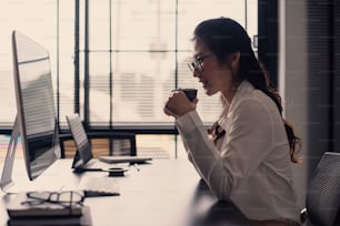 Charmante employée de bureau asiatique travaillant sur un ordinateur portable et appréciant de boire un café dans un bureau moderne.