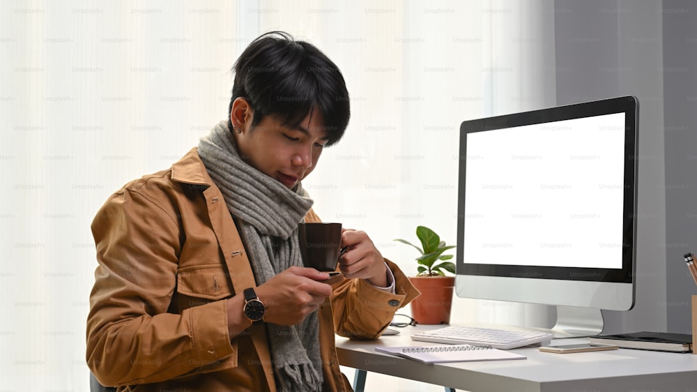 Attractive young man holding cup of to beverage and sitting in front of computer at home office.