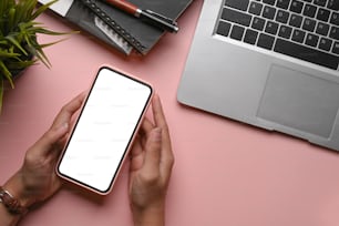 Overhead shot of female holding mock up smart phone with empty screen on pink desk.