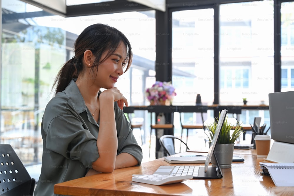 Vista lateral do designer da jovem mulher sentada no local de trabalho criativo e olhando para a tela do computador tablet.