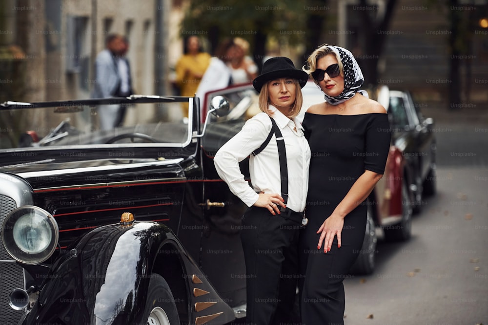 Women in old fashioned clothes stands together near retro car.