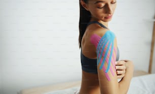 Sportive woman sits indoors with kinesio tape on her shoulder.