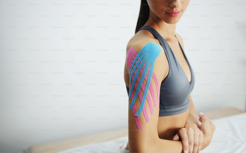 Sportive woman sits indoors with kinesio tape on her shoulder.