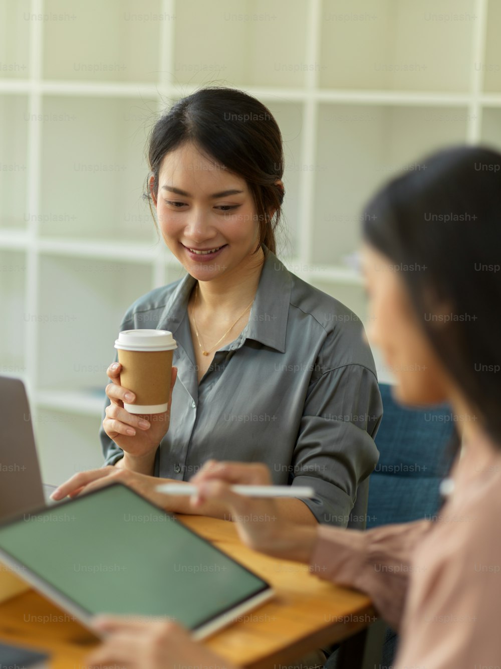 Portrait of female worker smiling and holding paper cup while listening her coworker present her idea