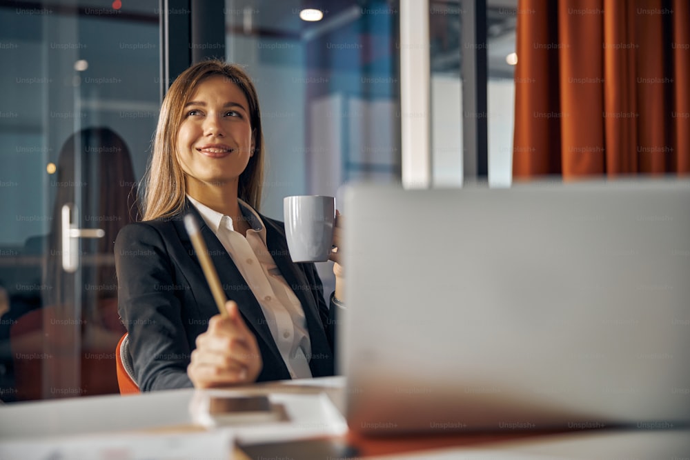 Waist-up portrait of a smiling dreamy female employee with a pencil and a mug of beverage looking away