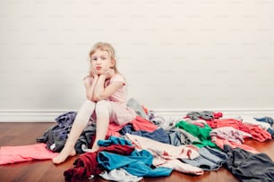 Mommy little helper. Bored Caucasian girl sorting clothes. Tired funny child arranging organazing clothing. Kid with messy stack of clothes things on floor. Home chores housework.