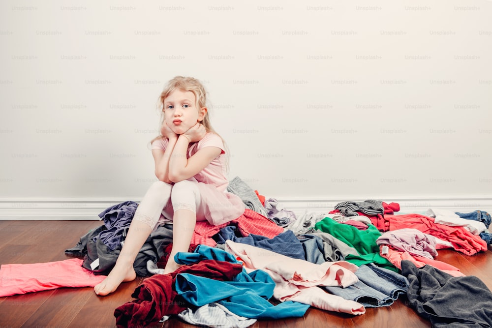 Mommy little helper. Bored Caucasian girl sorting clothes. Tired funny child arranging organazing clothing. Kid with messy stack of clothes things on floor. Home chores housework.