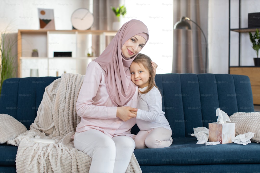Close up portrait of happy Muslim family, mother and little daughter, sitting together on blue sofa and hugging, posing to camera on the background of cozy light home interior. Motherhood concept.