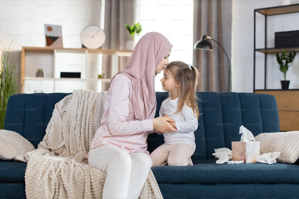 素敵な家族のお母さんと子供。ヒジャーブを着た幸せな愛情深い若いイスラム教徒の母親は、居心地の良いリビングルームのソファに座って、かわいい笑顔の小さな娘を抱きしめ、愛情を込めて額にキスをします。