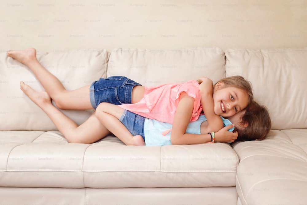 Two cute little Caucasian girls siblings playing at home. Adorable children kids lying hugging on couch together. Authentic candid lifestyle domestic life moment. Happy friends sisters relationship.