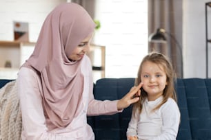 Close up of cute little girl child, sitting on sofa at home and smiling to camera, while her lovely pretty young Muslim mother in hijab applying moisturizing lotion or cream on her face. Skin care.
