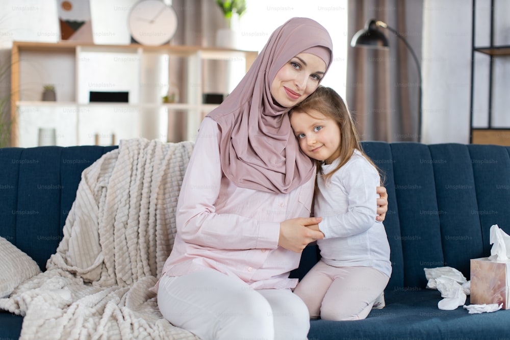 幸せなイスラム教徒の家族、母と小さな娘の肖像画を接写し、青いソファに一緒に座って抱きしめ、居心地の良い光の家のインテリアを背景にカメラに向かってポーズをとっています。母性の概念。