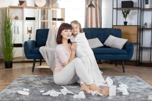 Child caring and hygiene routine. Pretty Caucasian mom hugs her little adorable 3 years old daughter after bath, wrapped in white towel, sitting together on carpet at stylish living room at home