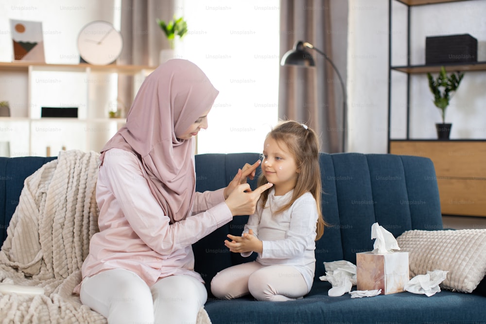 Happy Muslim woman mom and her little daughter sitting on the couch at home, having fun. Affectionate mother applying cream or lotion on face of her adorable kid girl.