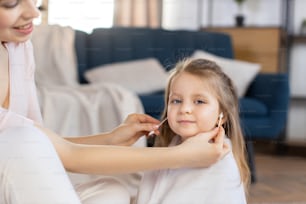 Close up de pequena menina loira de cabelos bonitos em toalha branca após o banho, sentada em sala aconchegante em casa, enquanto sua mãe carinhosa usa cotonetes e limpa as orelhas. Mamãe e menina em casa.