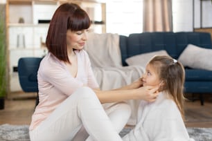 Beautiful Caucasian brunette affectionate mother uses cotton swabs and cleans the ears of small blond haired cute daughter in white towel, sitting in cozy room at home. Mom and little girl at home.
