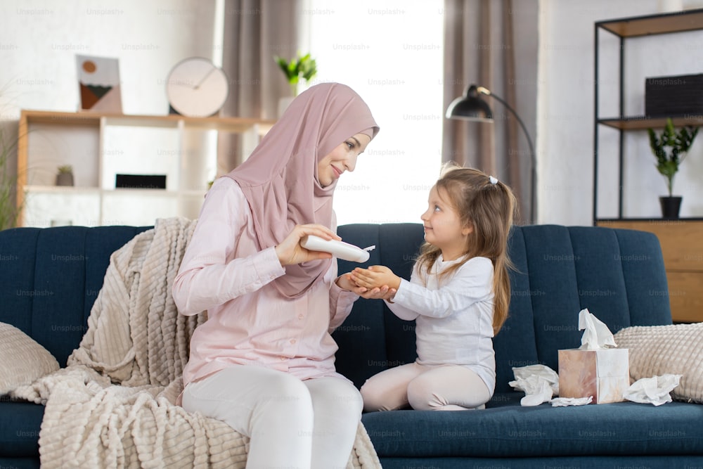 Indoor shot of lovely Muslim family doing hygiene routine at home. Young mom applying body cream or lotion on hands of little adorable daughter, looking each other.