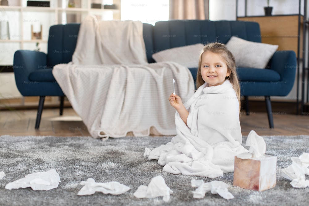 Portrait of little 3 years old cute blond girl, wrapped in towel after shower, holding hygienic cotton swab, sitting on the floor in light modern living room with blue sofa. Ear hygiene.
