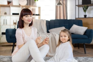 Young Caucasian mother holding hygienic cotton swabs, sitting on the floor in cozy living room with her cute little daughter, wrapped in towel after bathing at home. Hygiene and care concept.