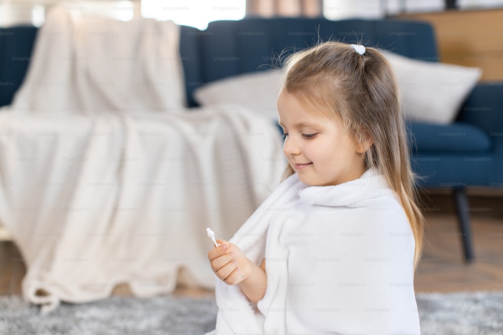 Ear cleaning, skincare concept. Little attractive kid girl, looking at cotton swab in her hand, sitting wrapped in towel after bath in modern appartment interior. Child's hygiene concept.