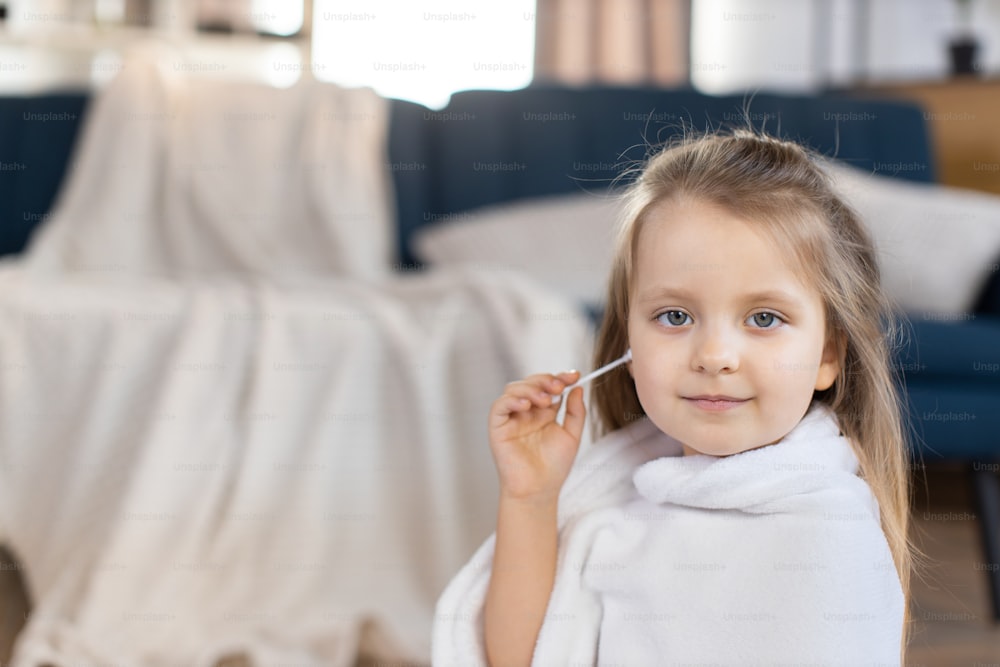 Nahaufnahme Innenporträt eines glücklich lächelnden dreijährigen Mädchens, das zu Hause vor der Kamera mit einem Lächeln posiert und ihr kleines Ohr mit Wattestäbchen reinigt. Hautpflege und Ohrenhygiene, Kopierraum.