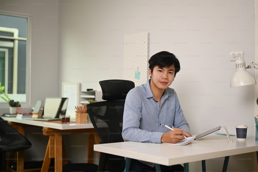 Empresário inteligente segurando tablet digital e sorrindo para a câmera enquanto está sentado em home office.