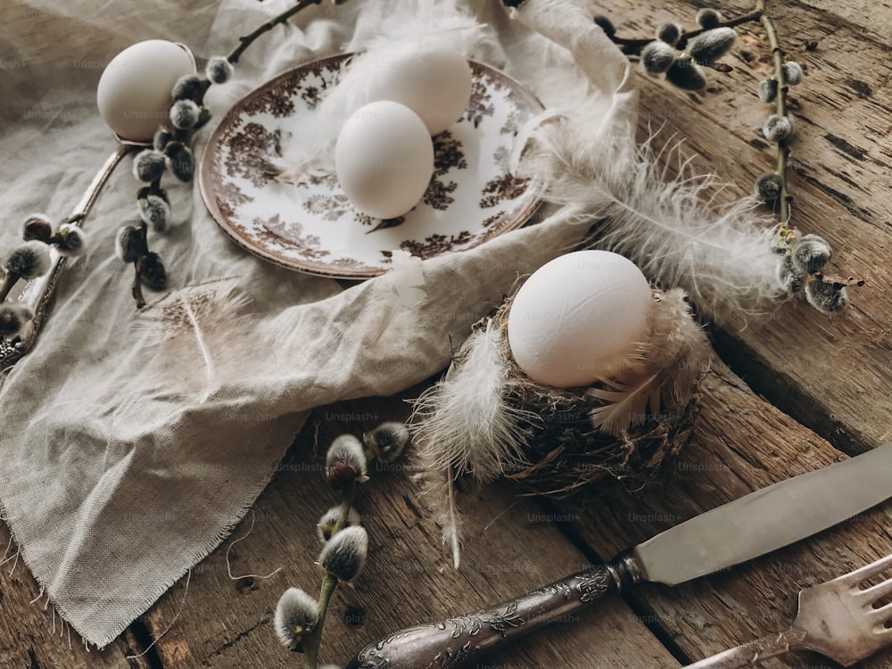Natural easter eggs, feathers, vintage plate and cutlery, napkin, pussy willow branches on aged wooden table. Happy Easter. Stylish rustic Easter table setting. Rural Easter still life