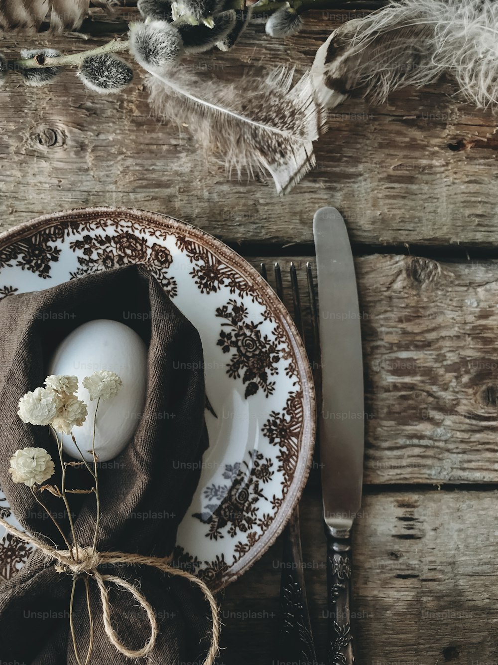 Stylish rustic Easter table setting. Natural easter egg in napkin with flowers, vintage plate and cutlery, soft feathers on aged wooden table. Rural Easter still life. Happy Easter