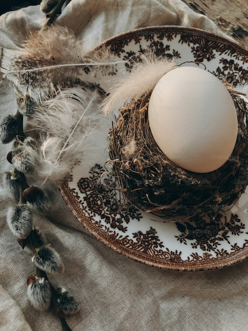 Natural easter egg in nest, vintage plate, soft feathers, linen napkin, pussy willow branches on aged wooden table.Stylish rustic Easter table setting.  Rural Easter still life