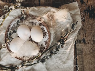 Natural Easter eggs on vintage plate, feathers, pussy willow branches on napkin on aged wooden table in sunny light. Rural Easter still life. Happy Easter. Stylish rustic table setting flat lay