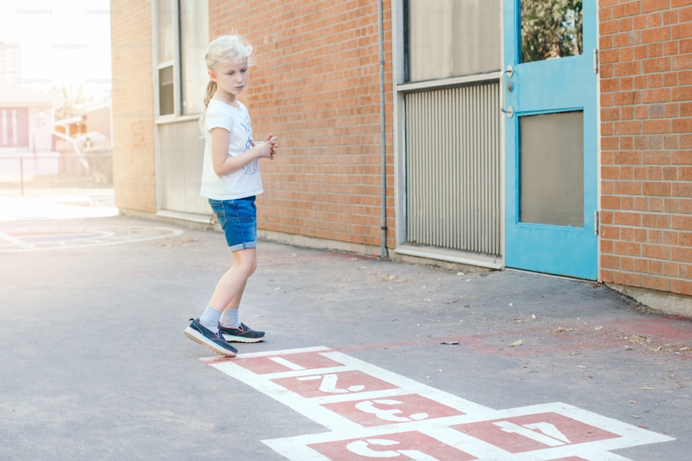 Young child girl playing jumping hopscotch on school yard outdoor. Funny activity game for kids on playground. Street sport for children. Happy childhood lifestyle.