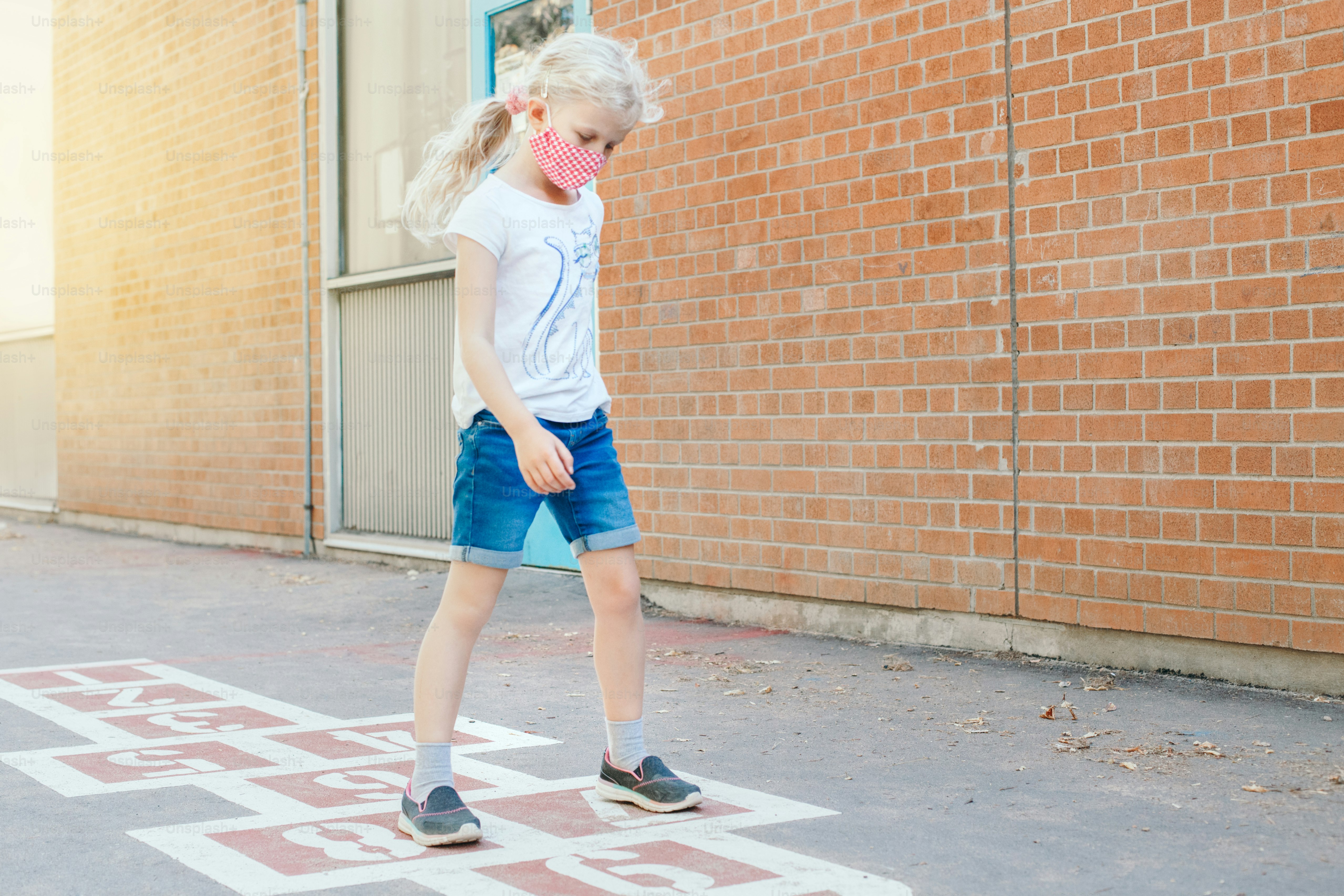 Child girl in face mask playing jumping hopscotch on school yard. Funny activity game for kids on playground outdoor. Street sport for children. Coronavirus covid-19 safety measures. New normal.