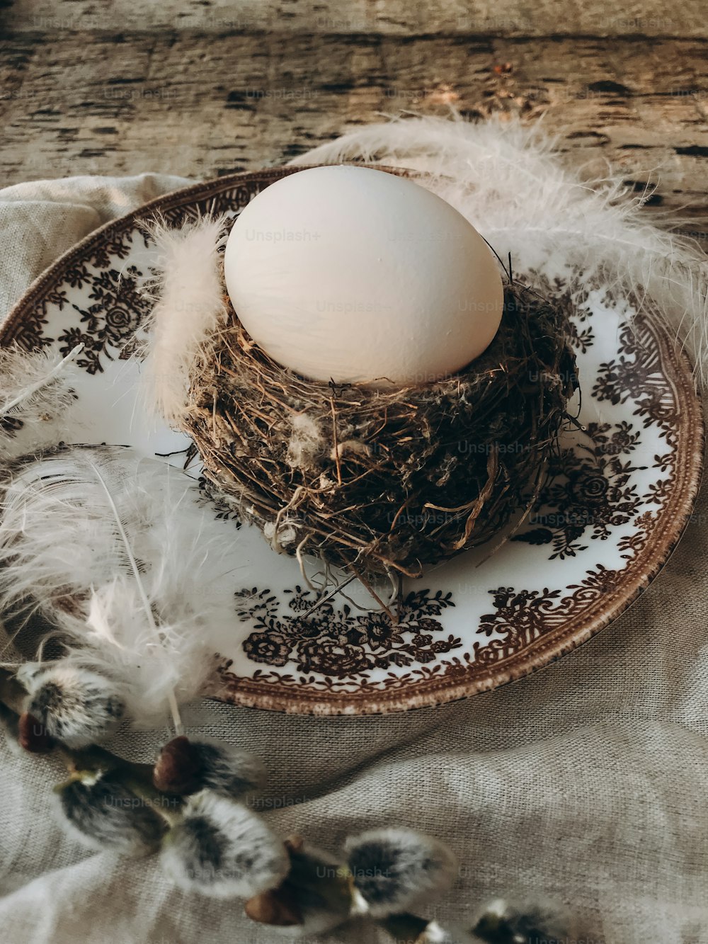 세련된 소박한 부활절 테이블 세팅. 빈티지 접시, 린넨 냅킨, 오래된 나무에 음부 버드 나무 가지에 부드러운 깃털이 있는 둥지에 자연 부활절 달걀. 시골 부활절 정물. 행복한 부활절