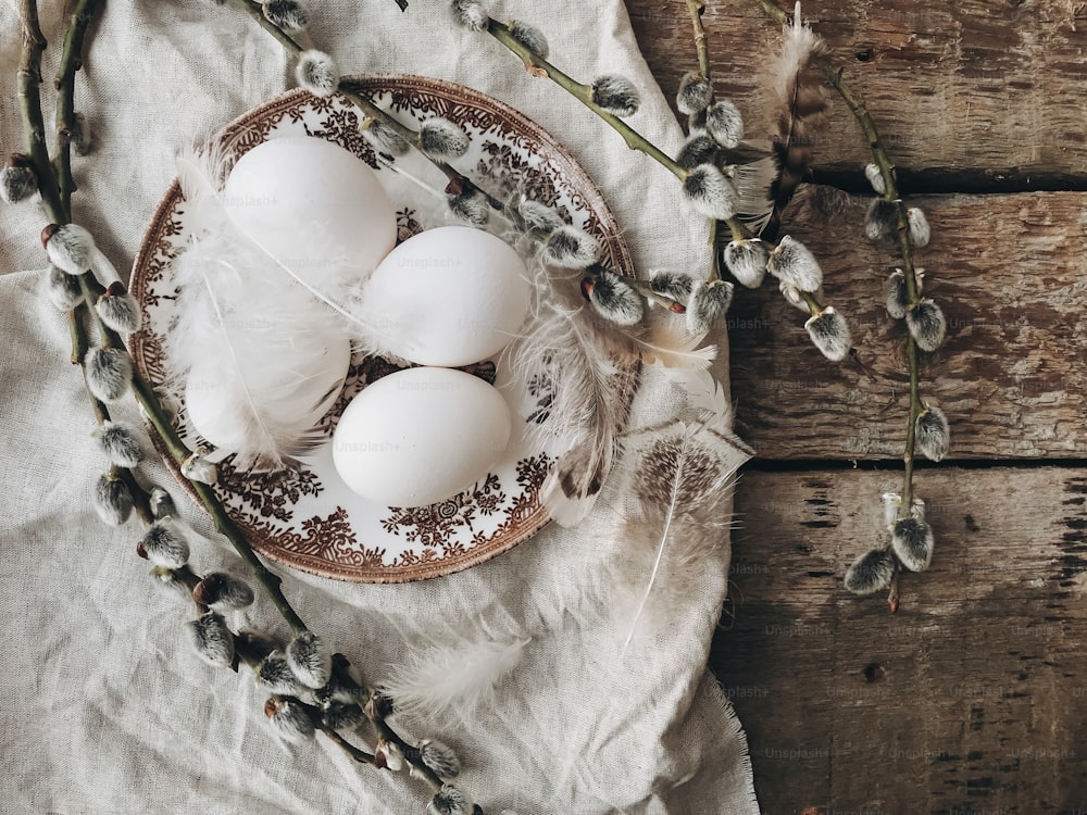 Stylish rustic Easter table setting flat lay. Natural easter eggs on vintage plate with feathers and pussy willow branches on napkin on aged wooden table. Rural Easter still life. Happy Easter