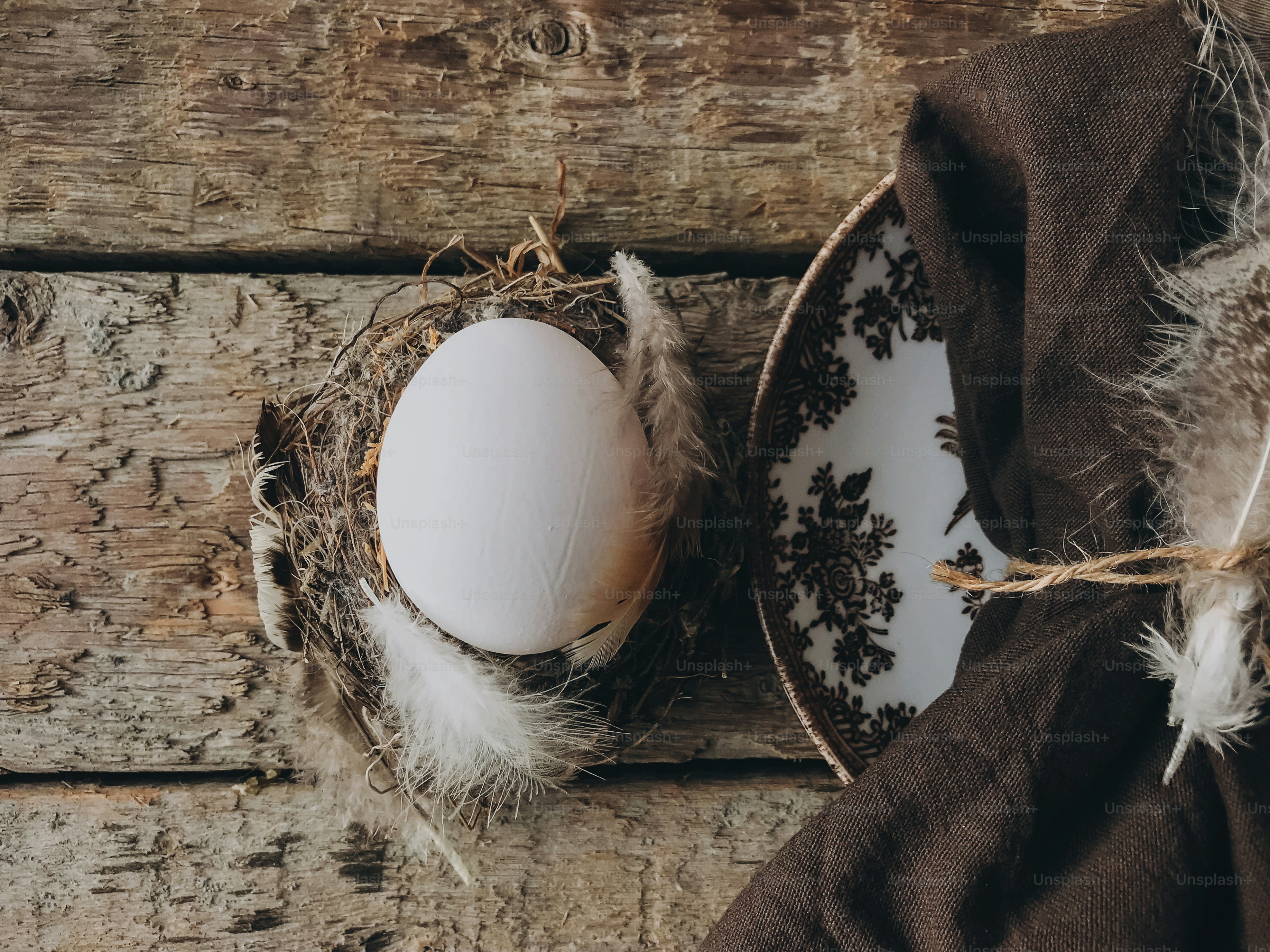 スタイリッシュで素朴なイースターテーブルセッティングフラットレイ。老朽化した木製のテーブルの上のビンテージプレートに羽とナプキンが入った巣の中の天然イースターエッグ。田舎のイースターの静物画。ハッピーイースター