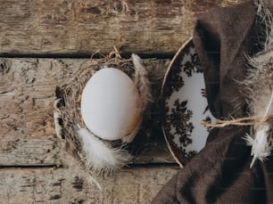 Stylish rustic Easter table setting flat lay. Natural easter egg in nest with feathers and napkin on vintage plate on aged wooden table. Rural Easter still life. Happy Easter