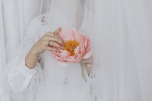 Beautiful stylish woman behind soft white tulle holding peony in hands. Young female gently holding big pink peony flower. Sensual soft image. Spring aesthetics. Womens day