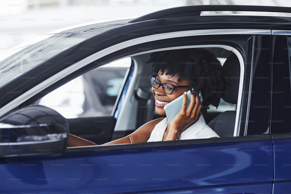 Converse por telefone. Mulher afro-americana jovem senta-se dentro do carro moderno novo.