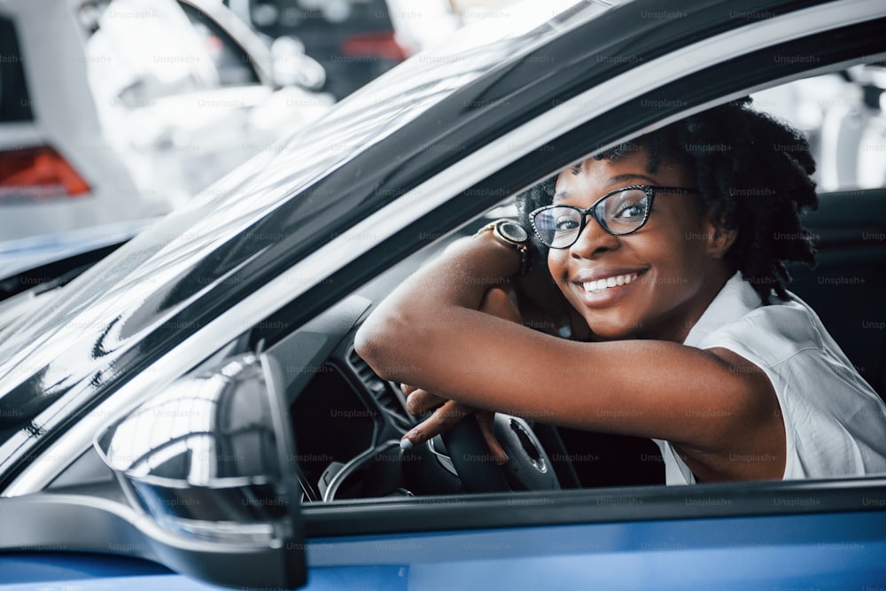 Cheerful smile. Young african american woman sits inside of new modern car.