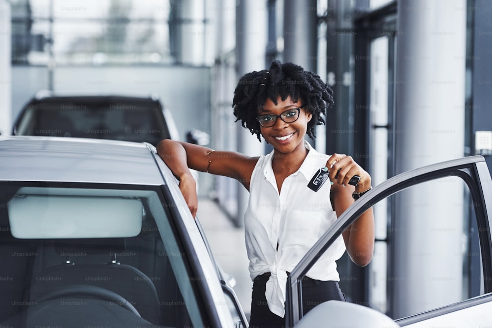 Jovem mulher afro-americana de óculos fica no salão de carros perto do veículo com as chaves nas mãos.