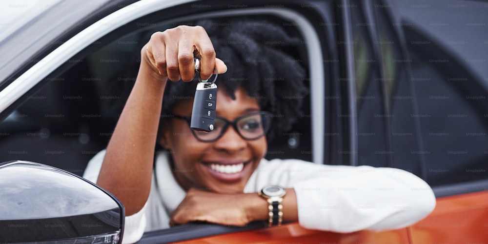 Young african american woman sits inside of new modern car and shows keys.