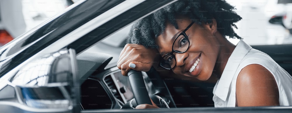 Cheerful smile. Young african american woman sits inside of new modern car.