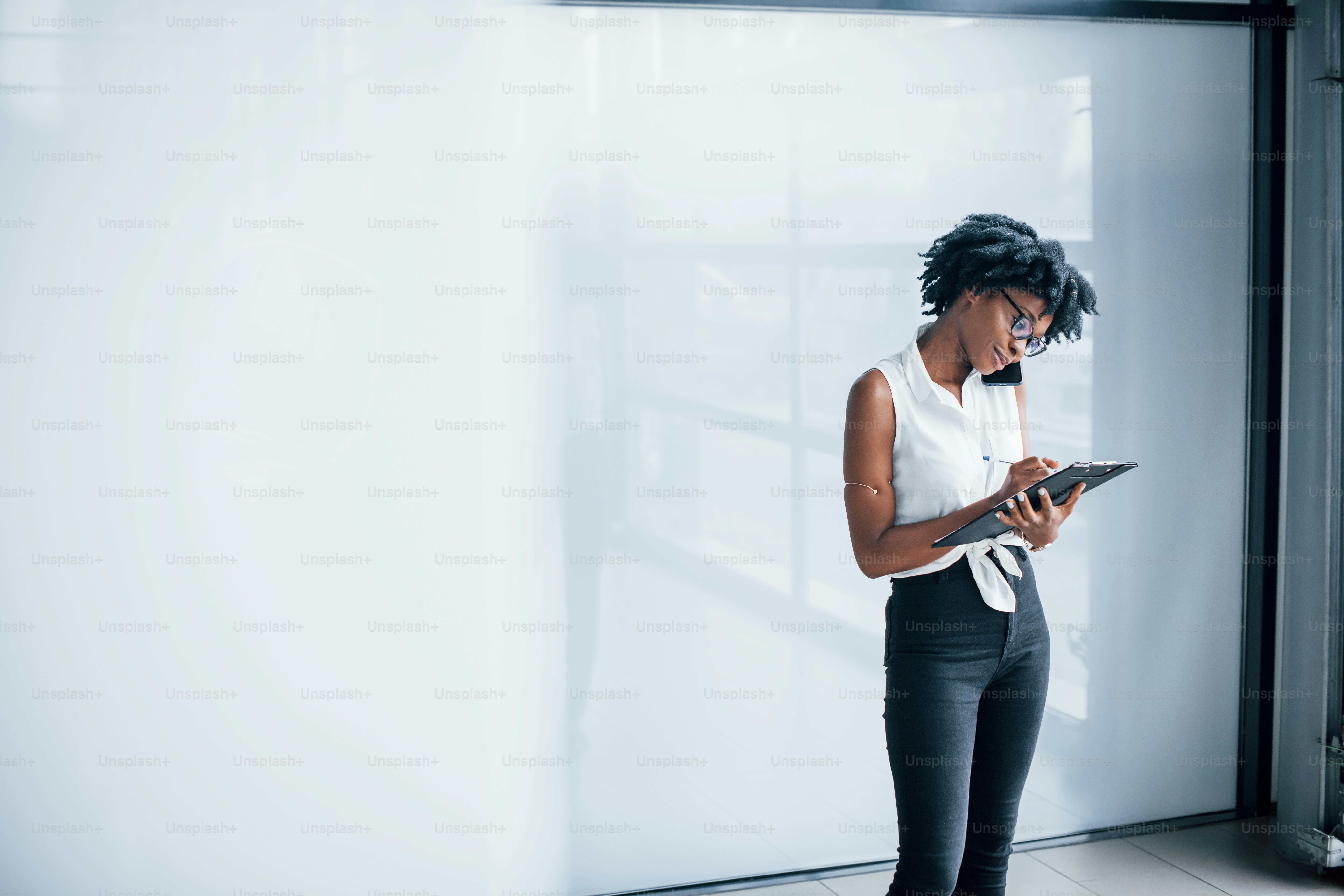 Young african american woman in glasses stands indoors in the office with tablet in hands