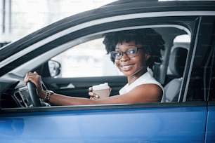 With cup of drink. Young african american woman sits inside of new modern car.