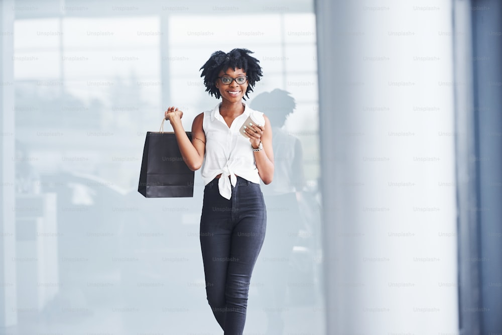 Young african american woman in glasses stands indoors with package in hands.