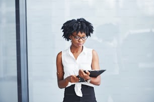 Young african american woman in glasses with notepad in hands stands indoors.